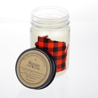 White candle contained in a clear glass jar with black tin lid propped on the side. A red and black plaid cut-out shape of Wisconsin on the face of the jar. 