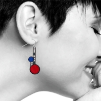 Polished brass dangle earring with asymetrical design by Frank Lloyd Wright featuring lower red circle and smaller blue circle above. Shown dangling from woman's ear. 