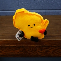 Yellow, soft plush toy in the shape of Wisconsin "sits" on the edge of a brown table. The stuffed state shape has a face, two small legs, and two short arms.