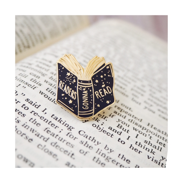 Metal lapel pin in shape of book with dark blue enamel and gold design. The pin is resting on a page in a book.